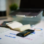 Smartphone showing a calculator app on a desk beside printed financial charts, a notebook, a keyboard and a laptop