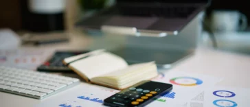 Smartphone showing a calculator app on a desk beside printed financial charts, a notebook, a keyboard and a laptop