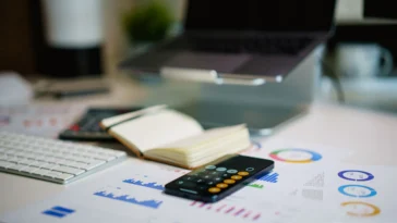 Smartphone showing a calculator app on a desk beside printed financial charts, a notebook, a keyboard and a laptop