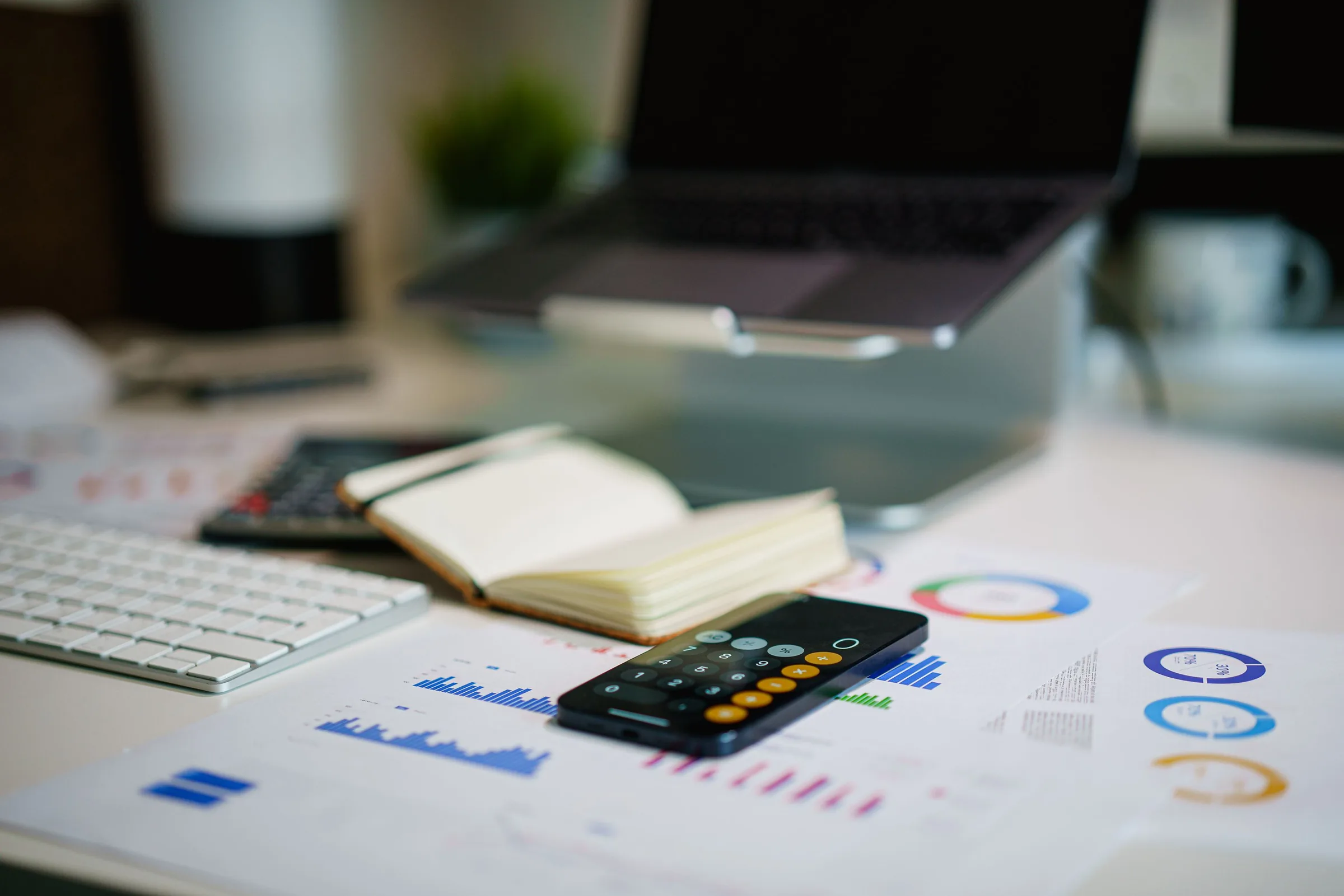 Smartphone showing a calculator app on a desk beside printed financial charts, a notebook, a keyboard and a laptop