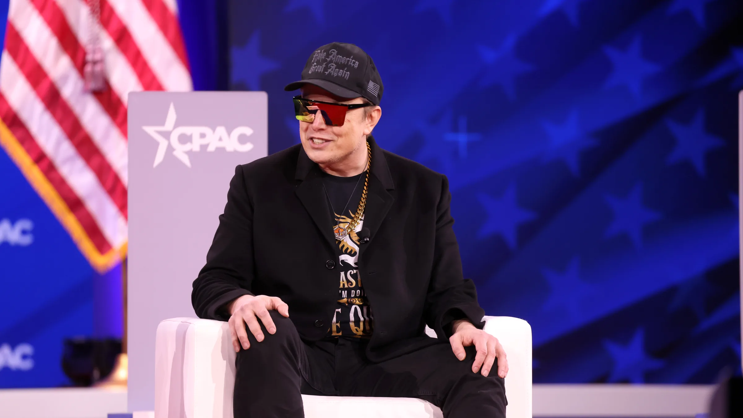 Elon Musk seated in a white armchair on stage at CPAC 2025, with a US flag and CPAC signage behind him