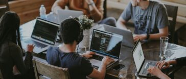 Five people collaborating on laptops around a wooden table in a coworking space