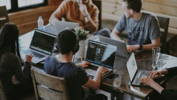 Five people collaborating on laptops around a wooden table in a coworking space