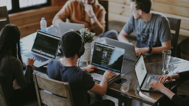 Five people collaborating on laptops around a wooden table in a coworking space