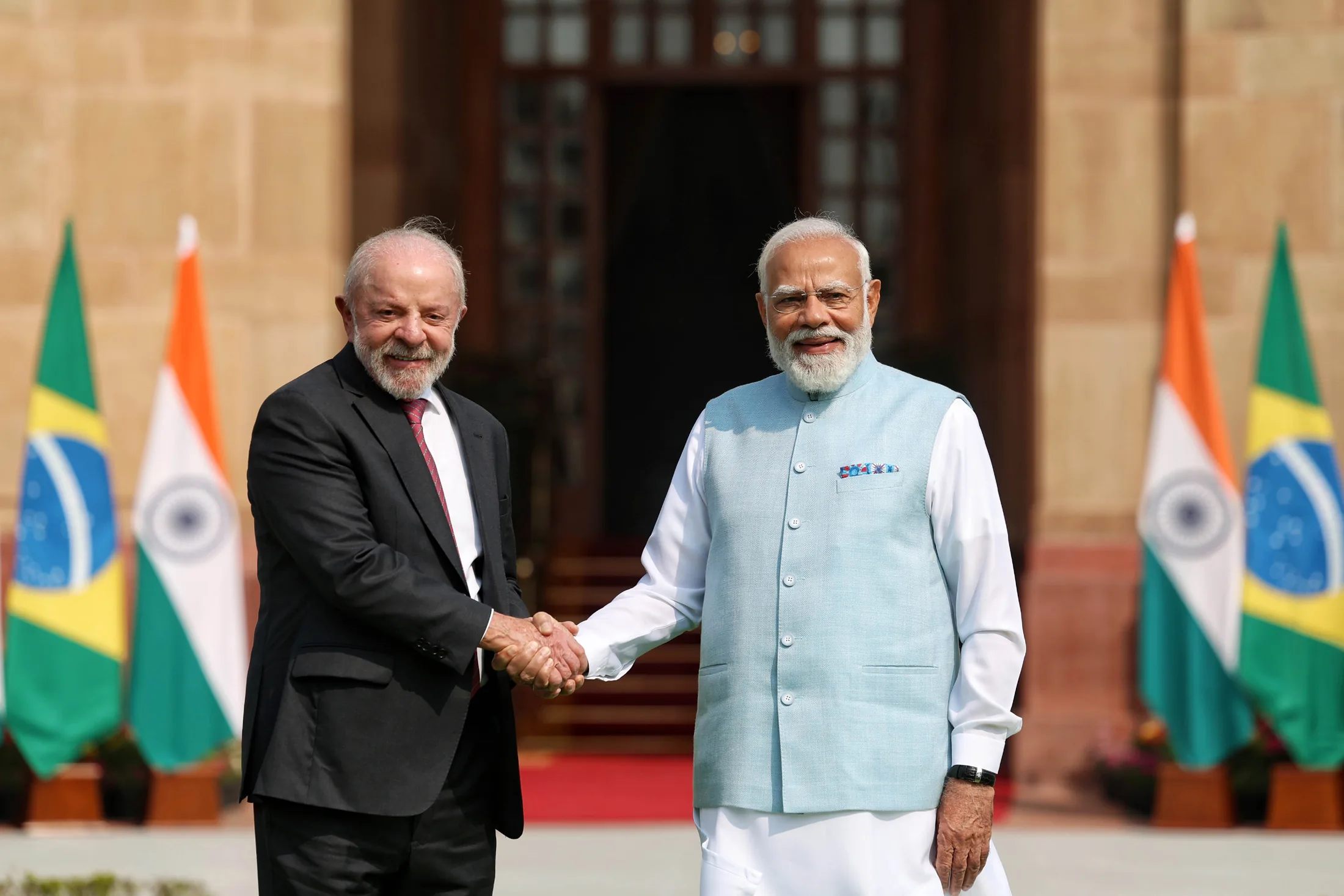 Prime Minister Narendra Modi and Brazilian President Luiz Inácio Lula da Silva shaking hands warmly in front of the Indian and Brazilian national flags at Hyderabad House in New Delhi