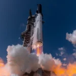 Starship's Super Heavy booster firing its ring of Raptor engines as it rises past the launch tower into a deep blue sky through orange-lit clouds of exhaust