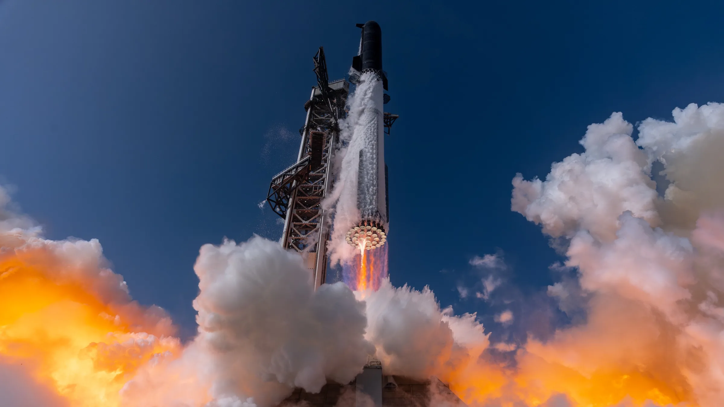 Starship's Super Heavy booster firing its ring of Raptor engines as it rises past the launch tower into a deep blue sky through orange-lit clouds of exhaust