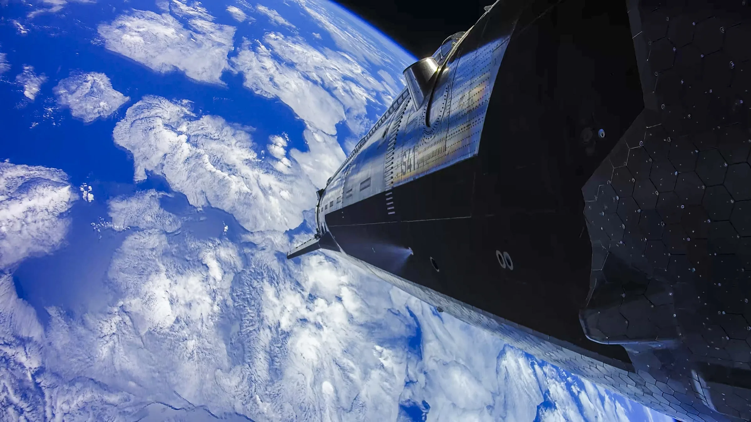 SpaceX Starship's dark steel hull seen from an onboard camera in orbit, with white clouds and blue ocean of Earth below