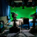 Two professional broadcast television cameras with teleprompters facing an anchor desk in a modern newsroom studio
