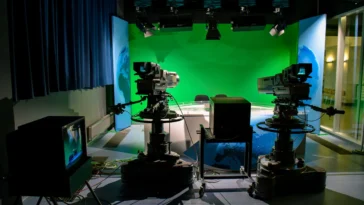 Two professional broadcast television cameras with teleprompters facing an anchor desk in a modern newsroom studio