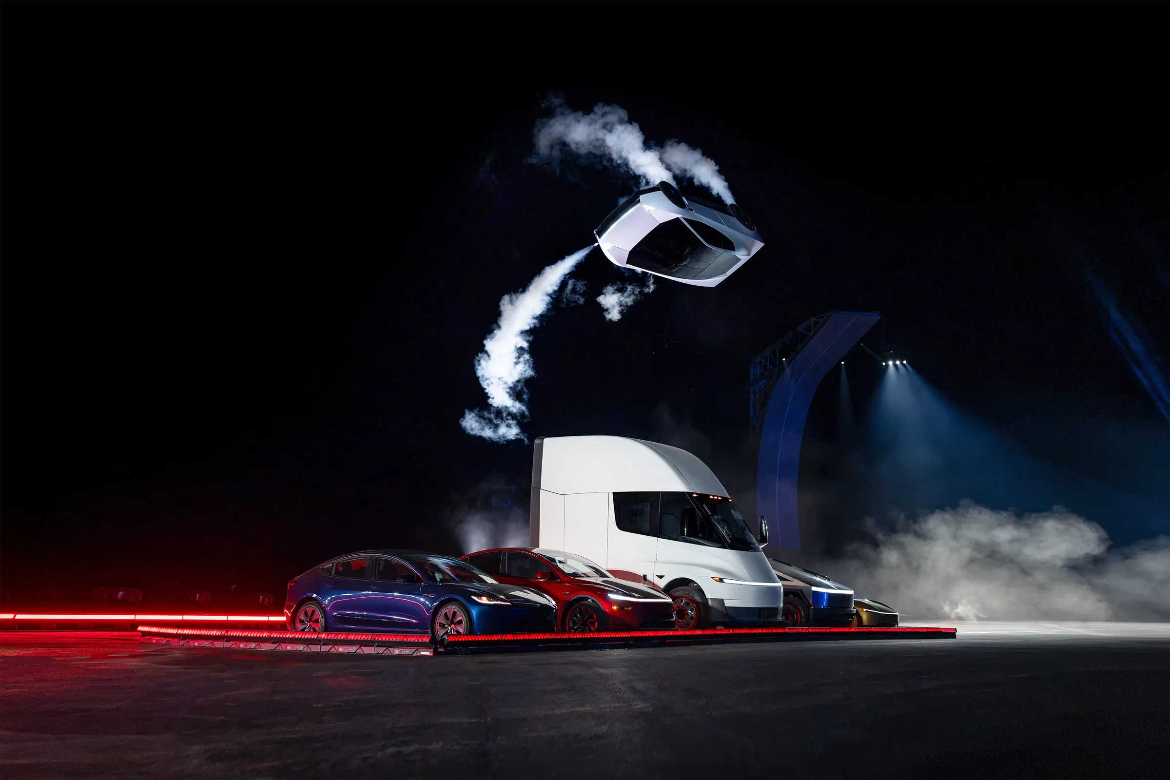 The next-generation Tesla Roadster flipping upside down through the air on cold-gas thrusters above a Tesla Semi and Model 3 and Model Y cars at night