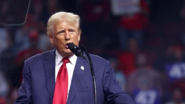 Donald Trump speaking at a podium during a campaign rally, with supporters in the stands behind him