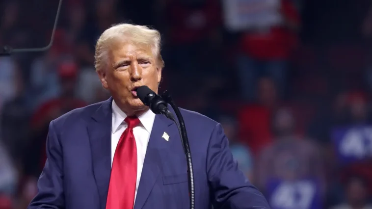 Donald Trump speaking at a podium during a campaign rally, with supporters in the stands behind him