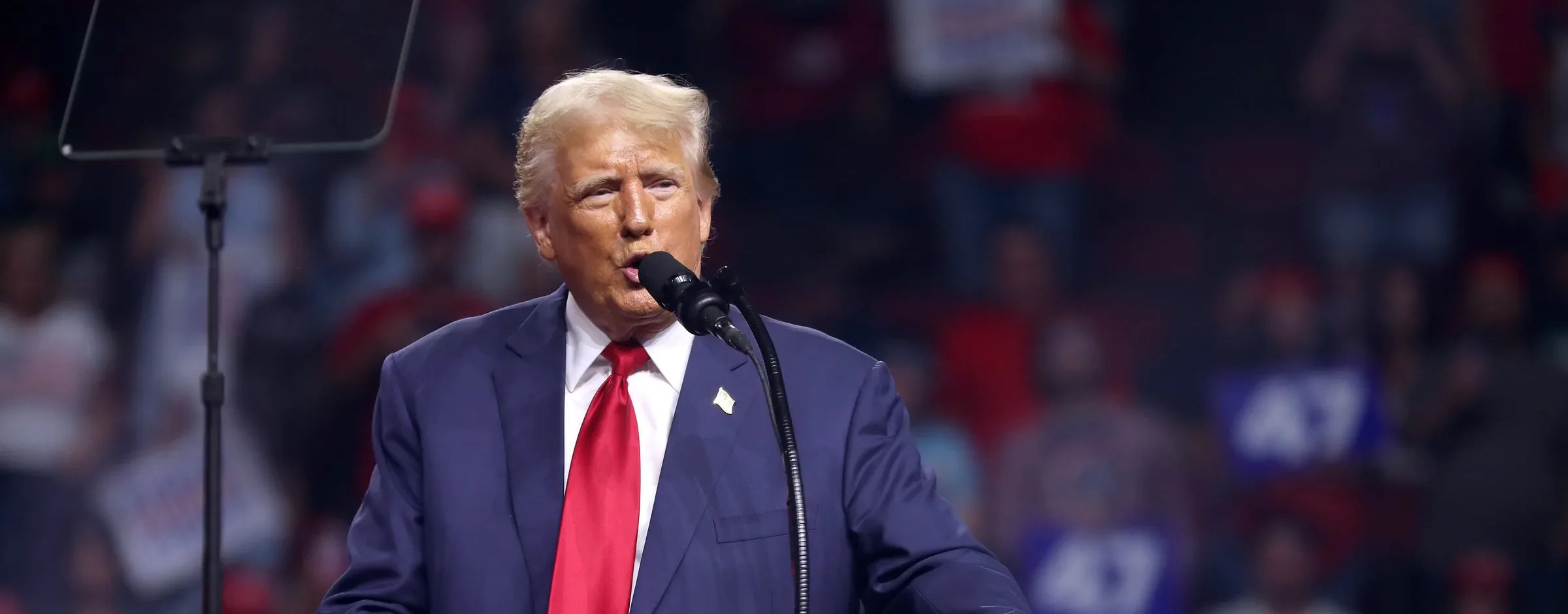 Donald Trump speaking at a podium during a campaign rally, with supporters in the stands behind him