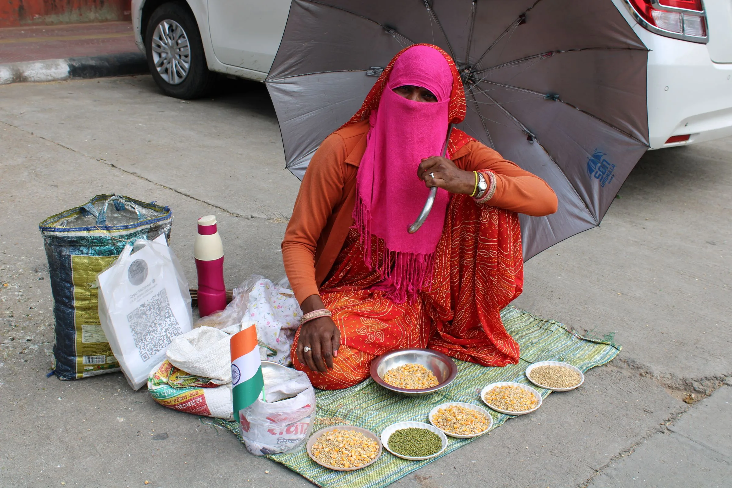 A street vendor in Jaipur sits under an umbrella beside bowls of grains, with a printed UPI QR code for payments on a bag beside her